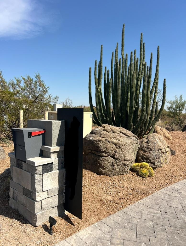 Desert landscaping with cacti, boulders, mailbox structure, and paved walkway
